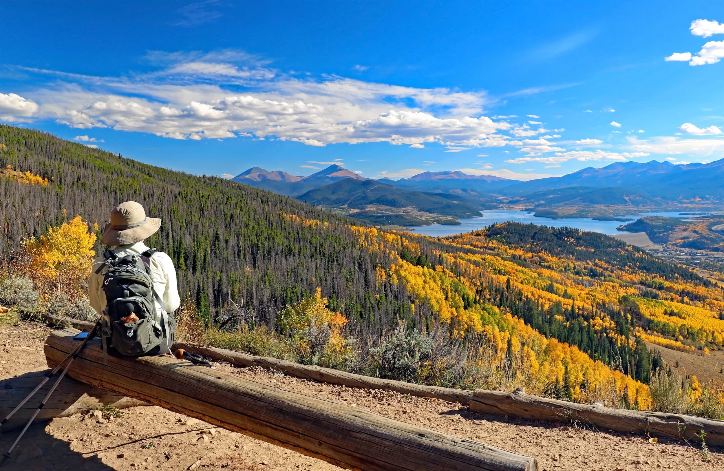 A hiker resting on a log bench above Dillon Reservoir, with aspens turning gold and peaks beyond
