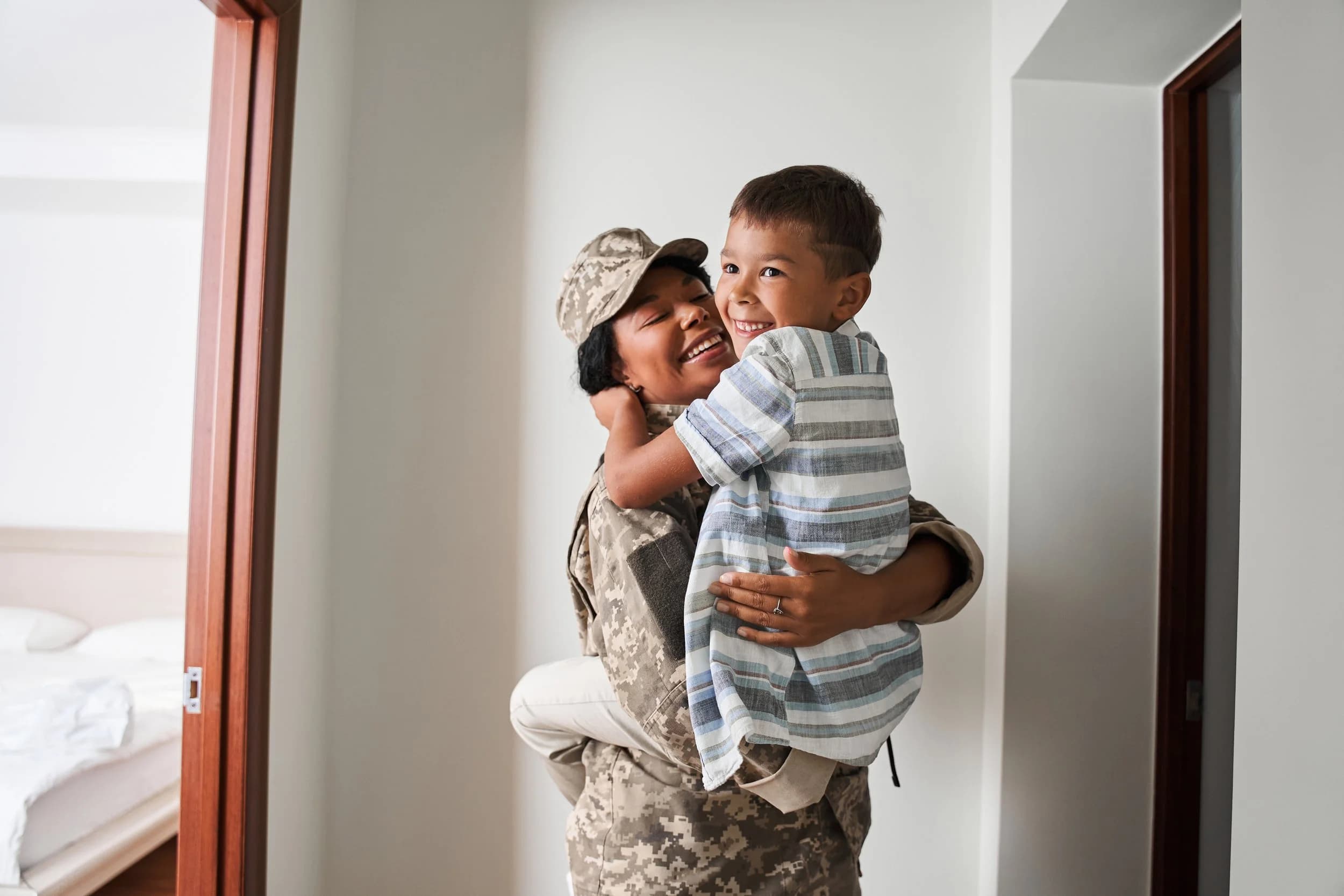 A mother in Army fatigues lifting her laughing young son in a hallway